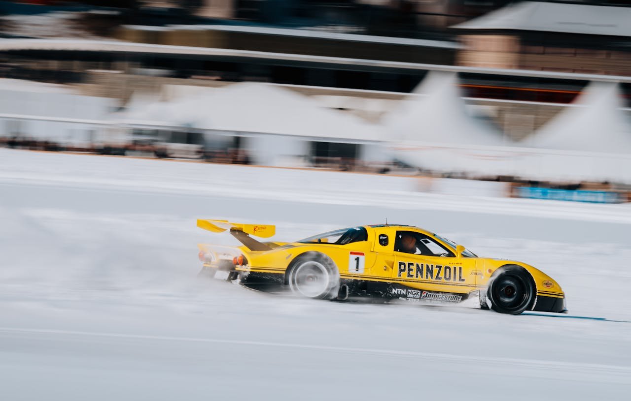 Yellow racing car with Pennzoil branding drifting on a snowy track during a winter motorsport event.
