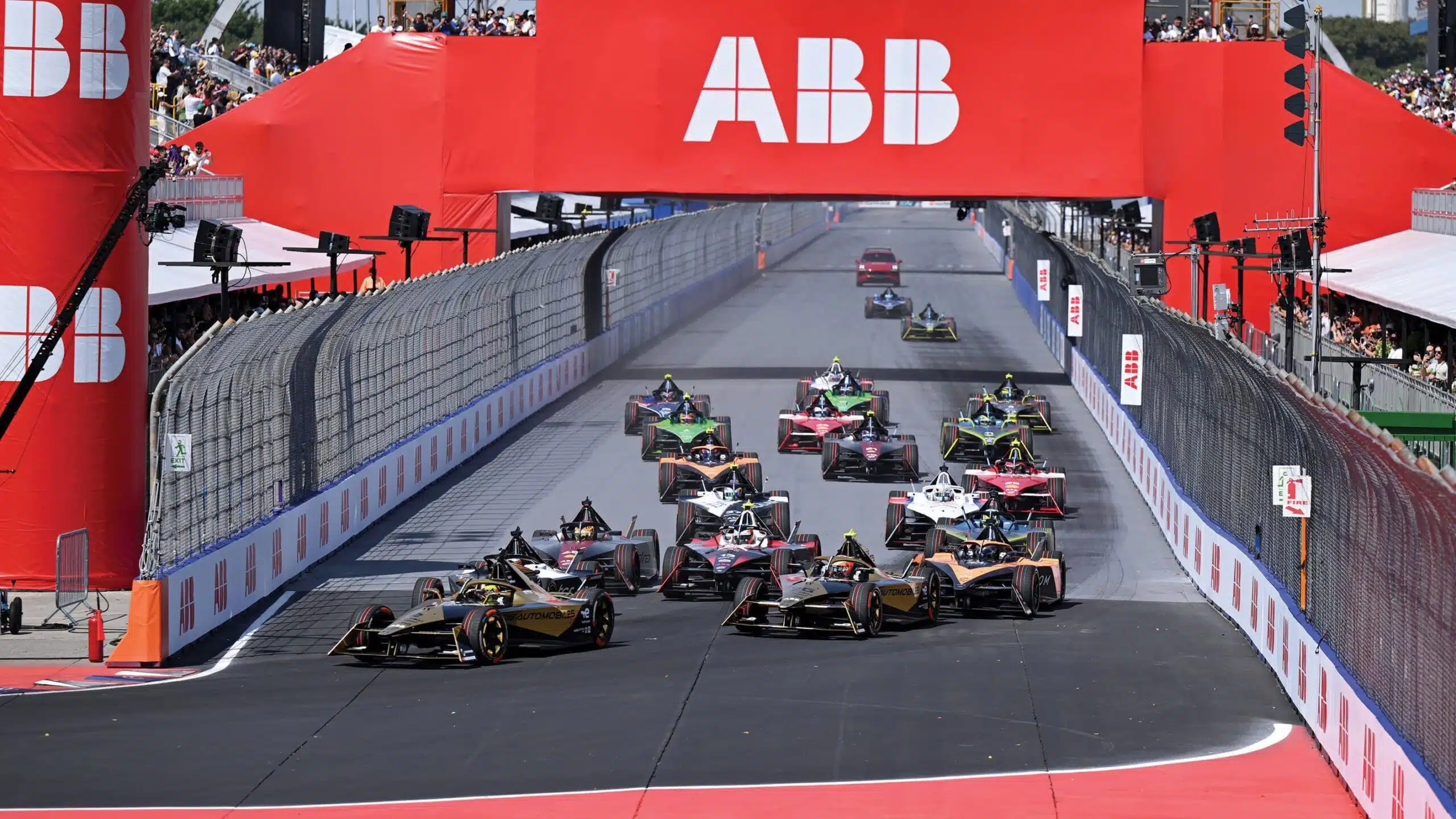 Formula E electric racing cars accelerating through a tight city street circuit corner during an E-Prix — narrow barriers, no run-off areas