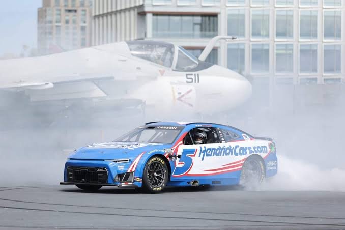 US Navy aircraft carrier docked at a naval base with city skyline in background, similar to the backdrop for NASCAR's Anduril 250 street race at Naval Base Coronado