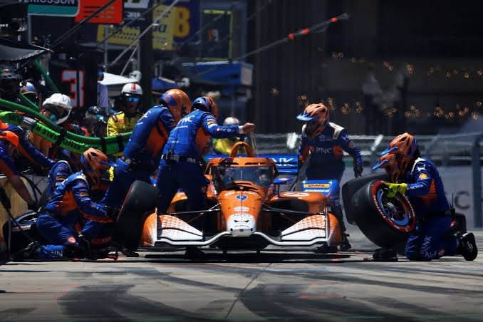 IndyCar pit lane with mechanics servicing cars — the hybrid system adds an energy management dimension to race strategy