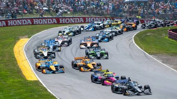 Open-wheel IndyCar racing through a tree-lined road course corner at speed